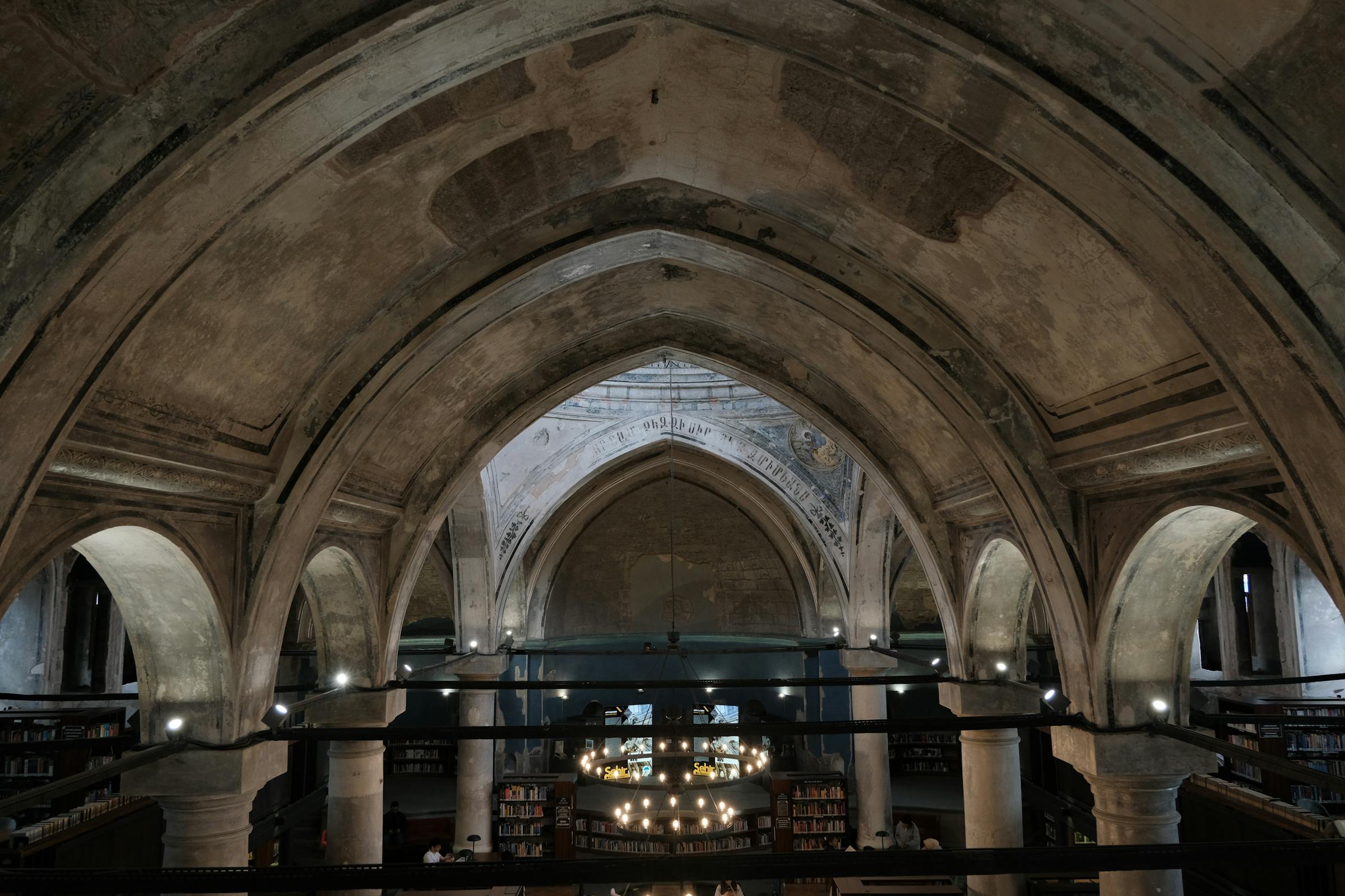 Interior of a vaulted reading room: ribbed gothic arches in stone, ranked above shelved volumes, the long silence of an institutional archive.